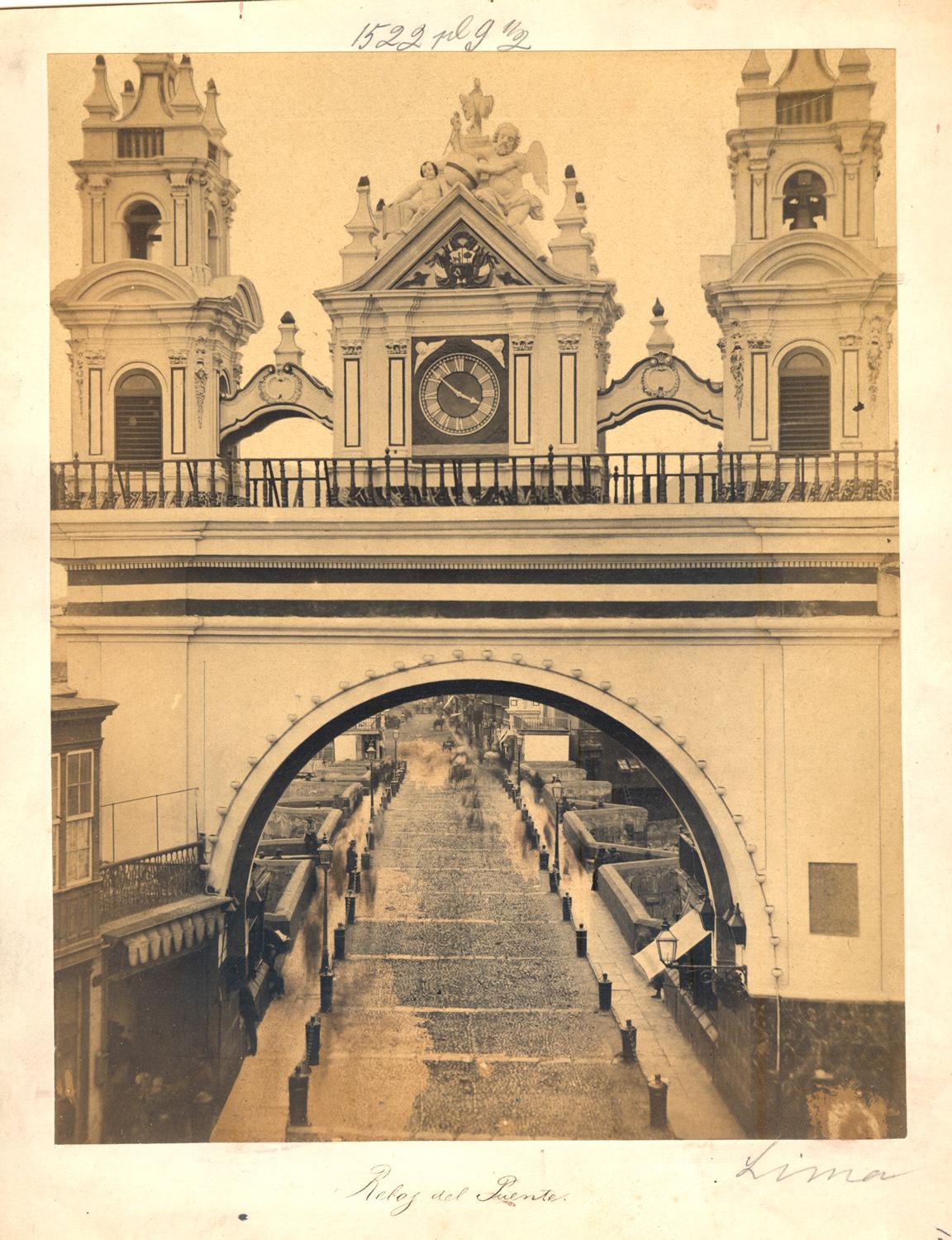 “Reloj del Puente”
Aquí vemos el desaparecido Arco del Puente que unía Lima con el Rímac. Éste marcaba el ingreso o salida a la ciudad. Este arco tenía la apariencia de una Iglesia, ya que contaba con dos torres como si se tratara del campanario. Hacia ambos lados y en la parte central, entre las dos torres se aprecia un reloj contenido en un elemento rectangular.Si agudizamos la vista cruzando el arco, el camino nos lleva por el histórico Puente de Piedra en dirección hacia el Rímac. El suelo se muestra empedrado y está dividido en 3 secciones: una central para la circulación de carretas y dos laterales que son las veredas para el tránsito de personas. Hacia ambos lados de la parte central y para que las carretas no invadan las veredas o sitio peatonal, se han colocado gruesas cadenas de acero sostenidas por antiguos cañones fijados (incrustados) verticalmente en el suelo. A estos elementos se les conocía como Guardacantones.
