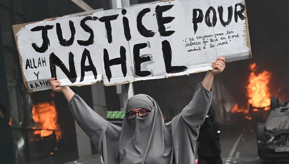 Una manifestante sostiene una pancarta que dice "Justicia para Nahel" mientras los autos son quemados en la calle en Nanterre, Francia. (Foto de Bertrand GUAY / AFP).