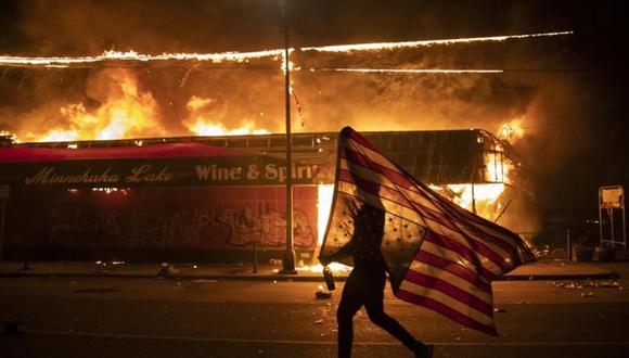 Un manifestante lleva una bandera estadounidense invertida frente a un edificio en llamas en Minneapolis. (Foto: AP/Julio Cortez).