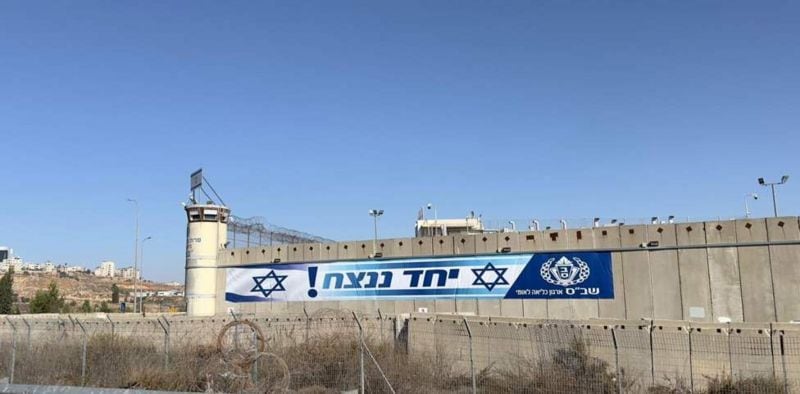 A banner hanging from an infrastructure with the phrase “United we will triumph!”  and the flag of Israel.