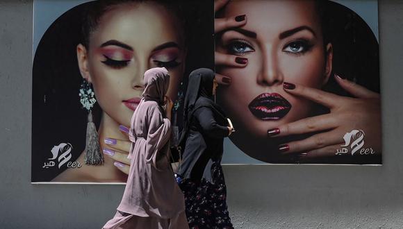 Mujeres vestidas con velo integral pasan junto a una valla publicitaria colocada en la pared de un salón de belleza en Kabul, Afganistán, el 7 de agosto de 2021. (SAJJAD HUSSAIN / AFP).