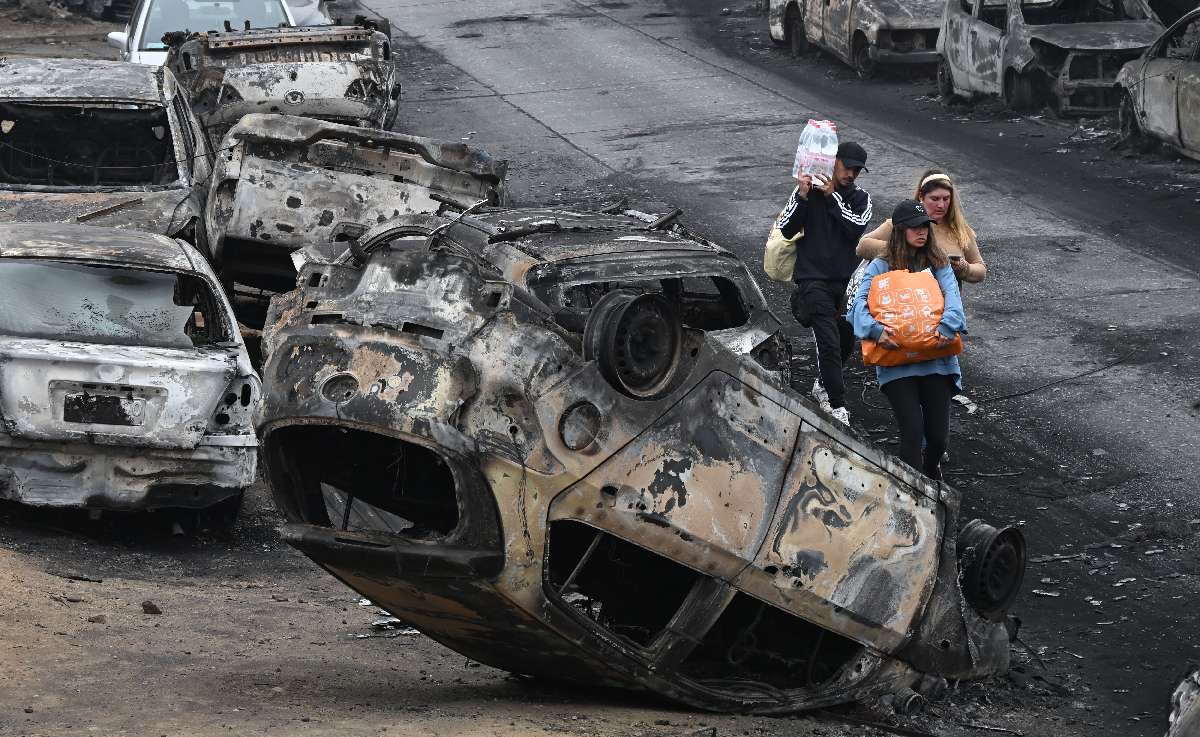 People walk past burned-out vehicles after a forest fire in Quilpue, Viña del Mar, Chile, on February 4, 2024. (Photo by RODRIGO ARANGUA/AFP).