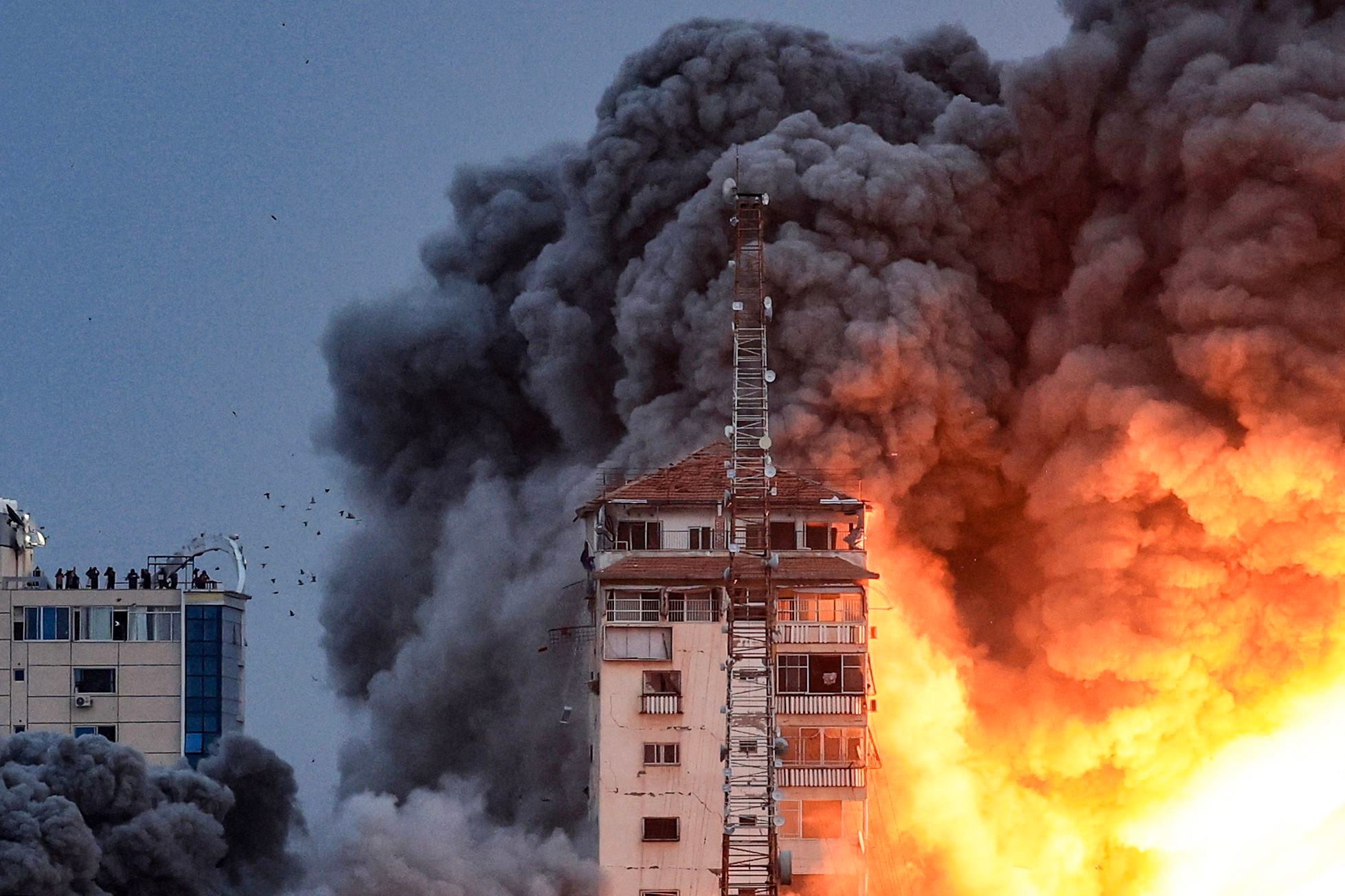 Personas paradas en una azotea observan cómo una bola de fuego y humo se eleva sobre un edificio en la ciudad de Gaza el 7 de octubre de 2023 durante un ataque aéreo israelí que golpeó el edificio de la Torre Palestina.(Foto de MAHMUD HAMS / AFP).
