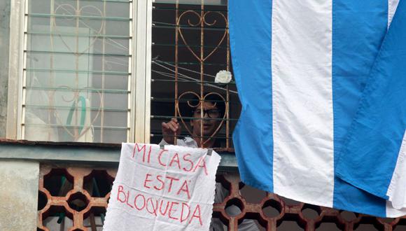 El disidente cubano y líder del movimiento Archipiélago Yunior García mira por la ventana de su departamento con un cartel que dice “Mi casa está bloqueada”, en La Habana, el 14 de noviembre de 2021. (AFP).