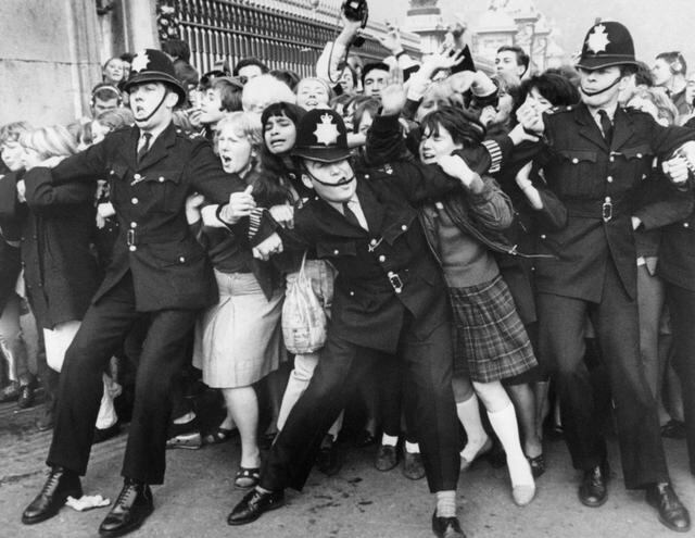 Un grupo de fans de The Beatles son retenidos por la policía en la cerca del Palacio de Buckingham, en el centro de Londres, durante una aparición pública del grupo cuando fueron recibidos por la reina Isabel II de Gran Bretaña. (Octubre de 1965-AFP)