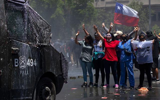 Un grupo de mujeres se para frente a un carro de la policía que reprime una marcha de trabajadores en Santiago de Chile. (Foto: AFP).