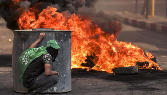 Un manifestante palestino enmascarado se esconde cerca de neumáticos en llamas durante los enfrentamientos con las fuerzas israelíes tras una manifestación de solidaridad con Gaza en la ciudad de Hebrón, en la Cisjordania ocupada. (Foto de HAZEM BADER / AFP).