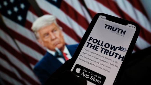 Esta foto ilustra a una persona que busca "Truth Social" en la tienda de aplicaciones de su teléfono inteligente, con una imagen del expresidente estadounidense Donald Trump en una pantalla de computadora de fondo. (CHRIS DELMAS / AFP).