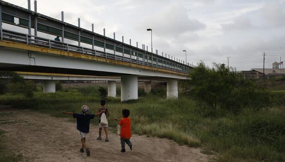 En esta fotografía de archivo del 26 de junio de 2019, niños migrantes caminan con sus familias a lo largo del Río Grande, mientras los peatones usan el puente Puerta México para ingresar a Brownsville, Texas. (AP/Rebecca Blackwell)