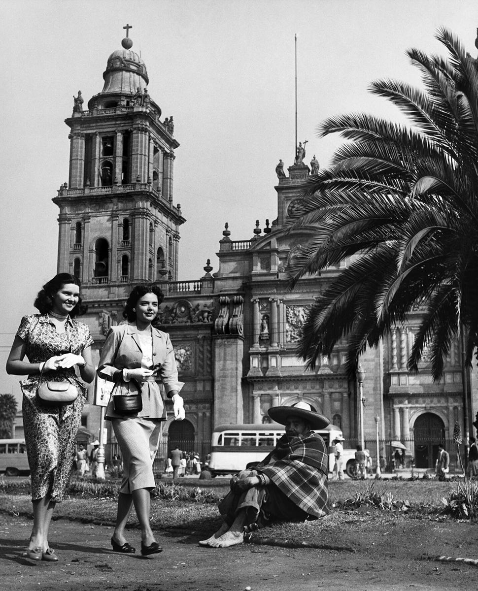 La señorita Heines y una acompañante pasean por el Zócalo después de visitar la catedral, de la serie "Turistas"
Ciudad de México, 20 de febrero de 1950. El reportero gráfico Juan Guzmán documentó la sociedad mexicana de mediados del siglo XX. Foto:
Colección y Archivo de Fundación Televisa