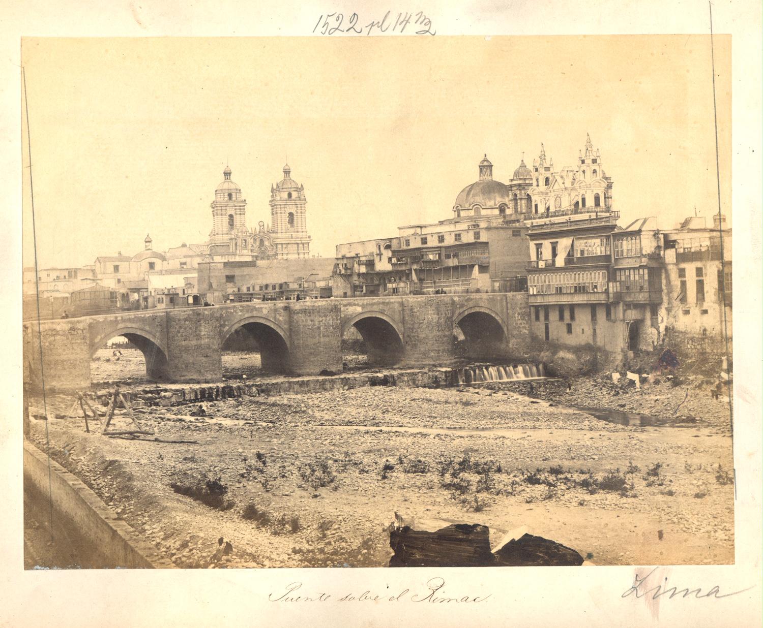 “Puente sobre el Rímac” (se ven torres de san francisco)
En esta foto tenemos una escena del Rio Rímac y el Puente de Piedra, vista desde el Rímac. Se ve en el lecho del río que corre un delgado hilo de agua. Se aprecia el Puente de Piedra, una construcción que data originalmente de la primera mitad del siglo XVII y que conservamos en la actualidad con sus modificaciones. Hacia la derecha, encontramos las torres del Arco del Puente, además de la cúpula y una de las torres de la desaparecida Iglesia de Desamparados. Vemos al fondo hacia la izquierda, las torres de la Iglesia de San Francisco y la Iglesia de la Soledad.
