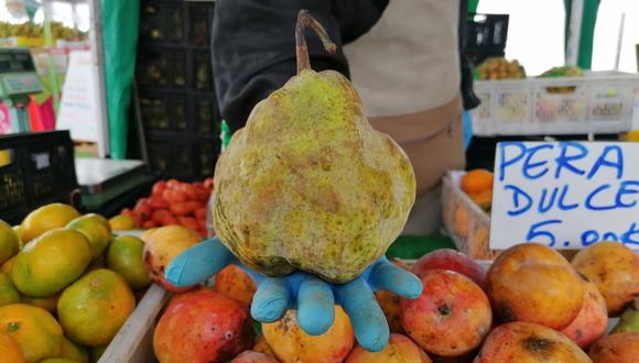 Un productor sostiene una pera dulce más grande que su propia mano que viene (junto a otras frutas) del norte chico. El puesto pertenece a la asociación de pequeños productores de Aguaymanto, Cajatambo.