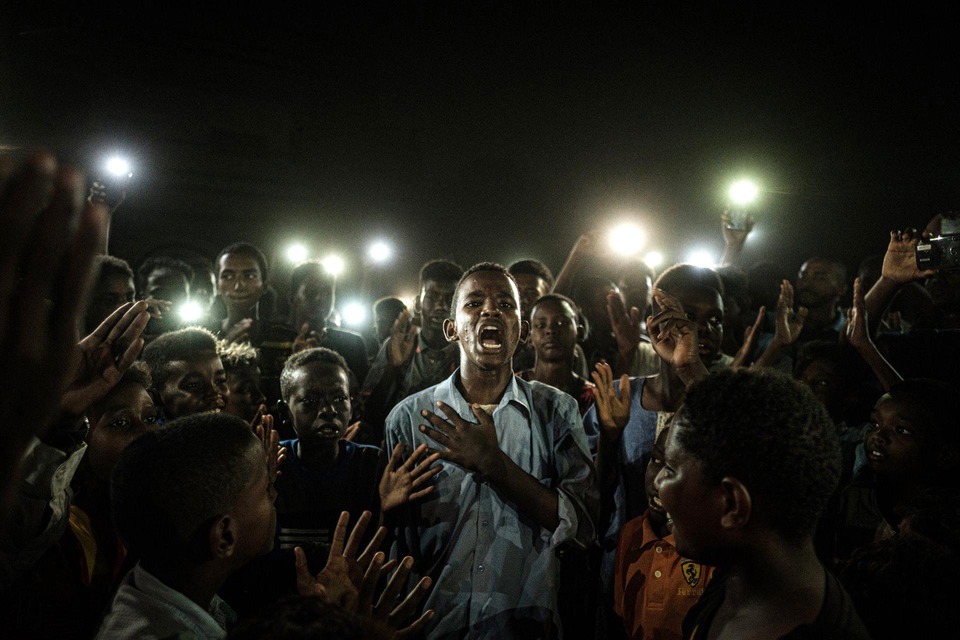 Foto ganadora. La gente canta consignas cuando un joven recita un poema, iluminado por teléfonos móviles, antes del diálogo directo de la oposición con la gente en Jartum el 19 de junio de 2019. (AFP / Yasuyoshi Chiba)