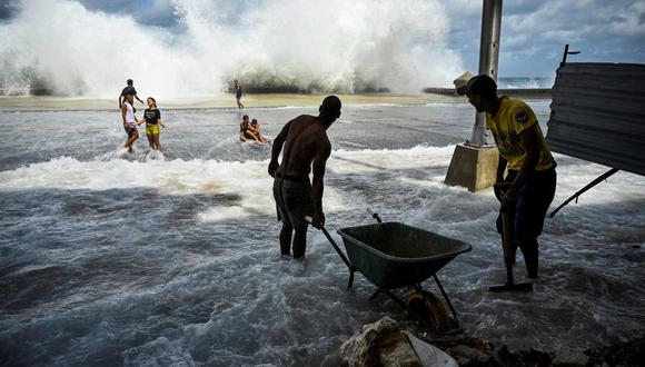 Trabajadores comunitarios recogen escombros mientras niños juegan cerca del Malecón de La Habana, el 29 de septiembre de 2022, tras el paso del huracán Ian. (Foto de YAMIL LAGE / AFP)