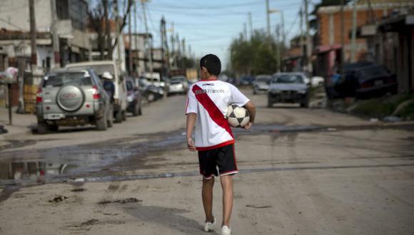 El niño argentino Daniel Dorado, que viste una camiseta del River Plate y sostiene una pelota de fútbol, ​​camina de regreso a casa, en el barrio pobre de Costa Eperanza, en la provincia de Buenos Aires. (Foto de ARIEL TIMY TORRES / AFP).