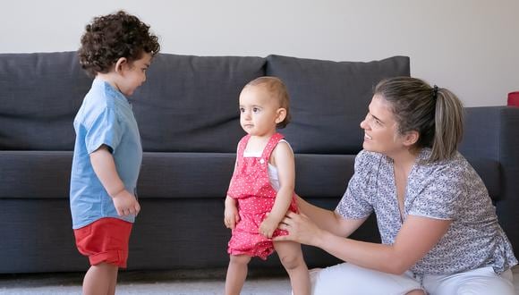 Happy mother playing with little children at home and sitting cross-legged. Cute baby girl and boy standing barefoot on carpet in living room. Family indoors, weekend and childhood concept