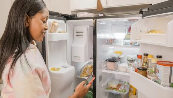 Una mujer mirando su celular mientras abre el refrigerador. | Imagen referencial: 
Kindel Media / Pexels