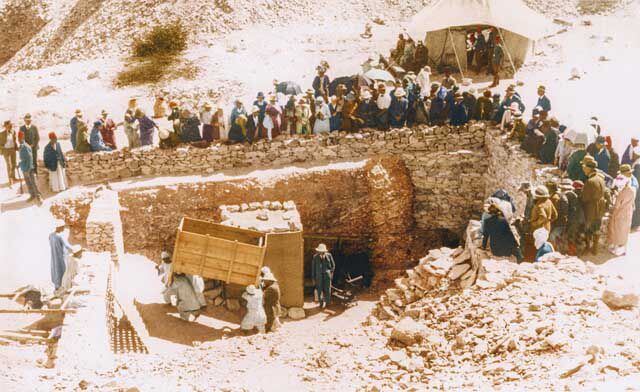 A crowd gathers at the gates of Tutankhamun's tomb as work crews remove a large object, possibly an armchair. 