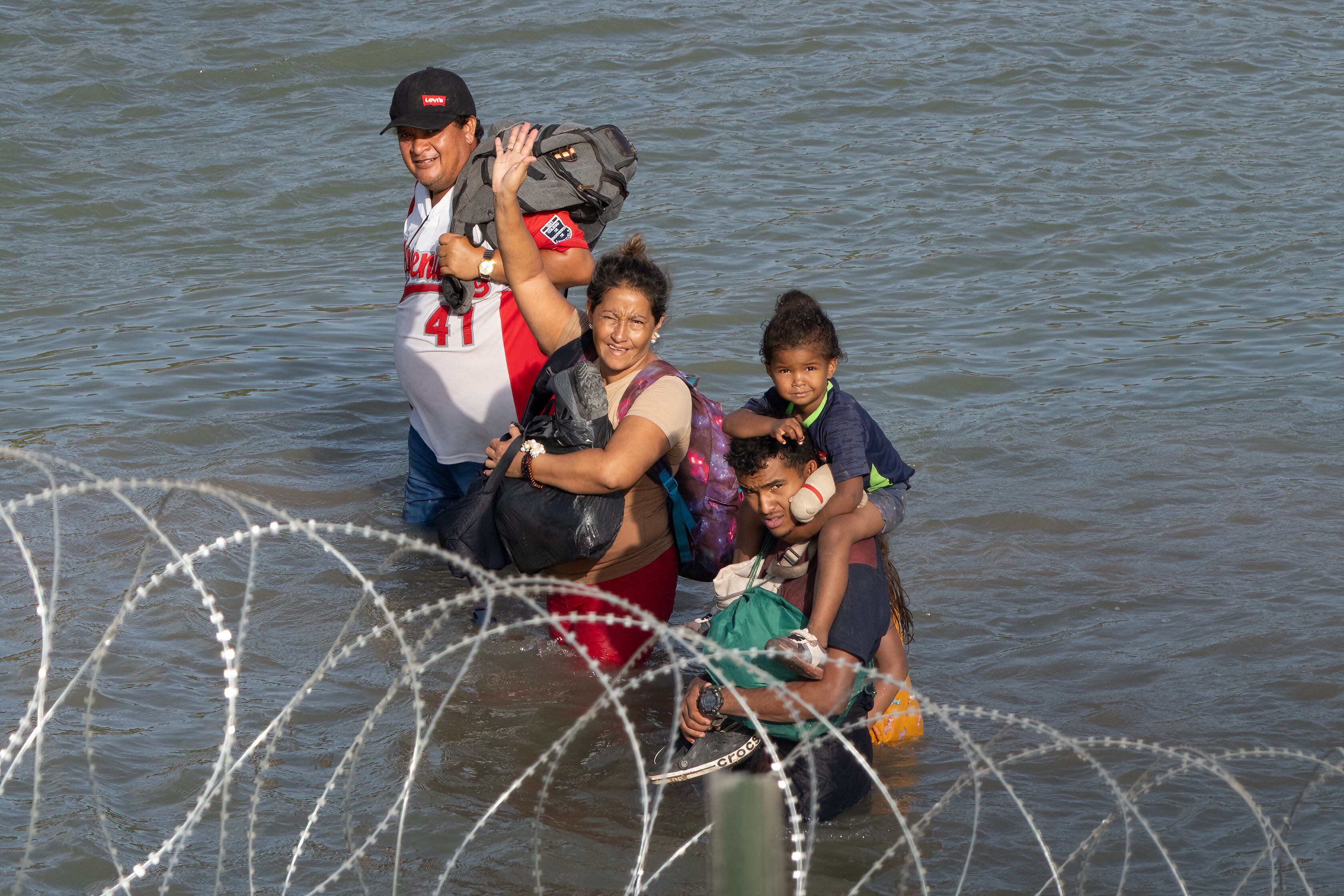 Migrantes saludan mientras caminan cerca de alambre de concertina en el agua a lo largo de la frontera del Río Grande con México. (Foto: AFP)