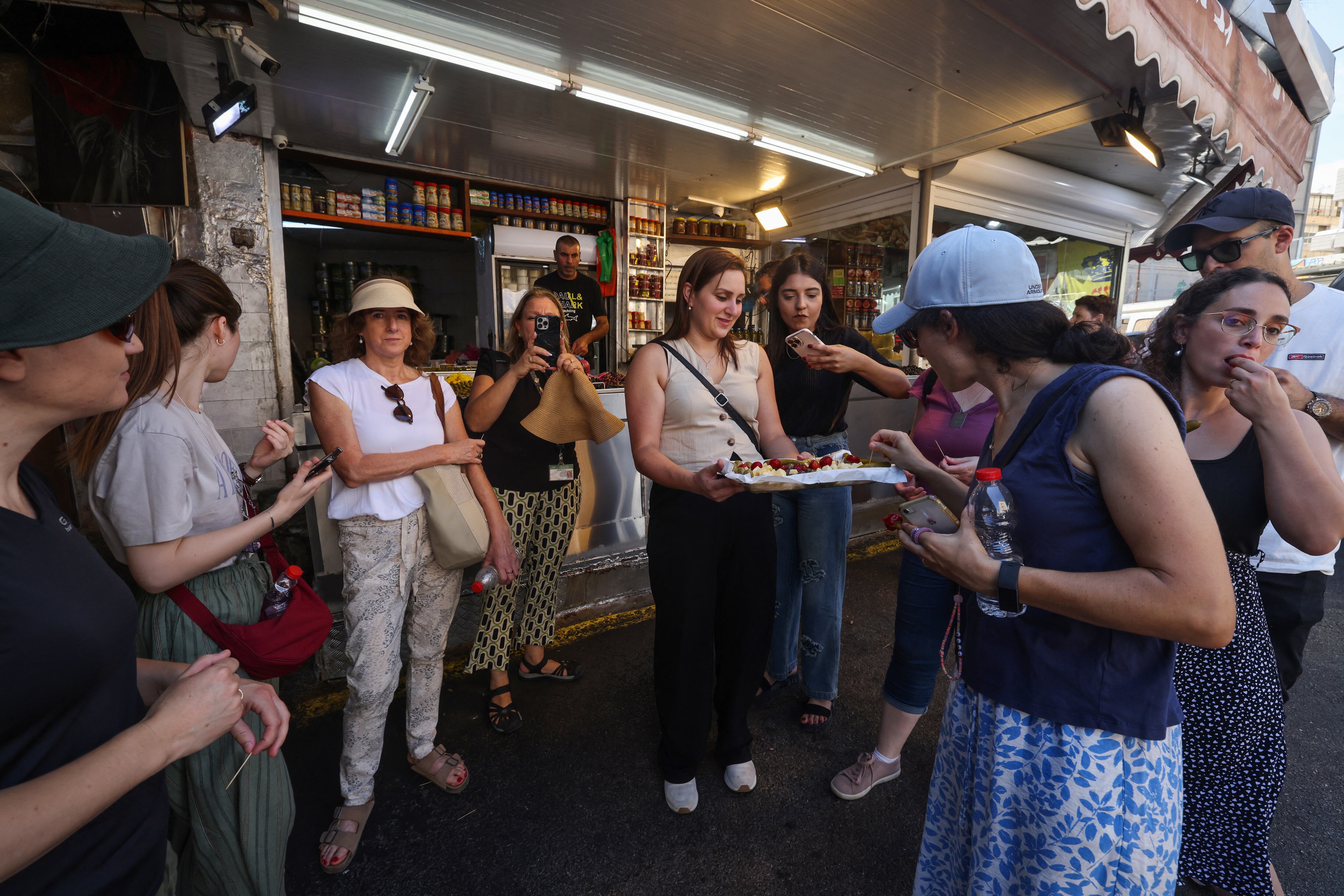 La gente prueba bocados de una bandeja de degustación ofrecida por una tienda en un mercado de la ciudad portuaria de Haifa, en el norte de Israel, el 5 de agosto de 2024. (Foto de Menahem KAHANA / AFP)
