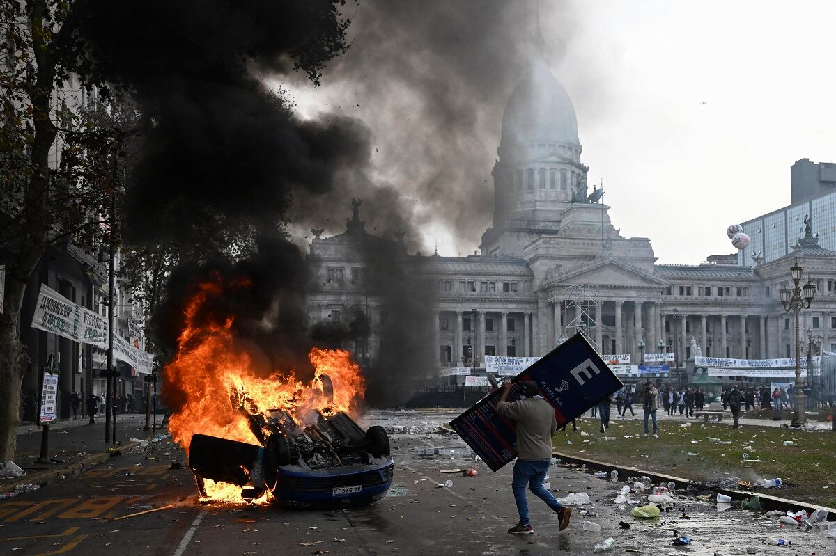 A protester with a sign approaches a Cadena 3 radio car that was set on fire during a protest in front of the National Congress, in Buenos Aires, on June 12, 2024. (Photo by Luis ROBAYO / AFP)