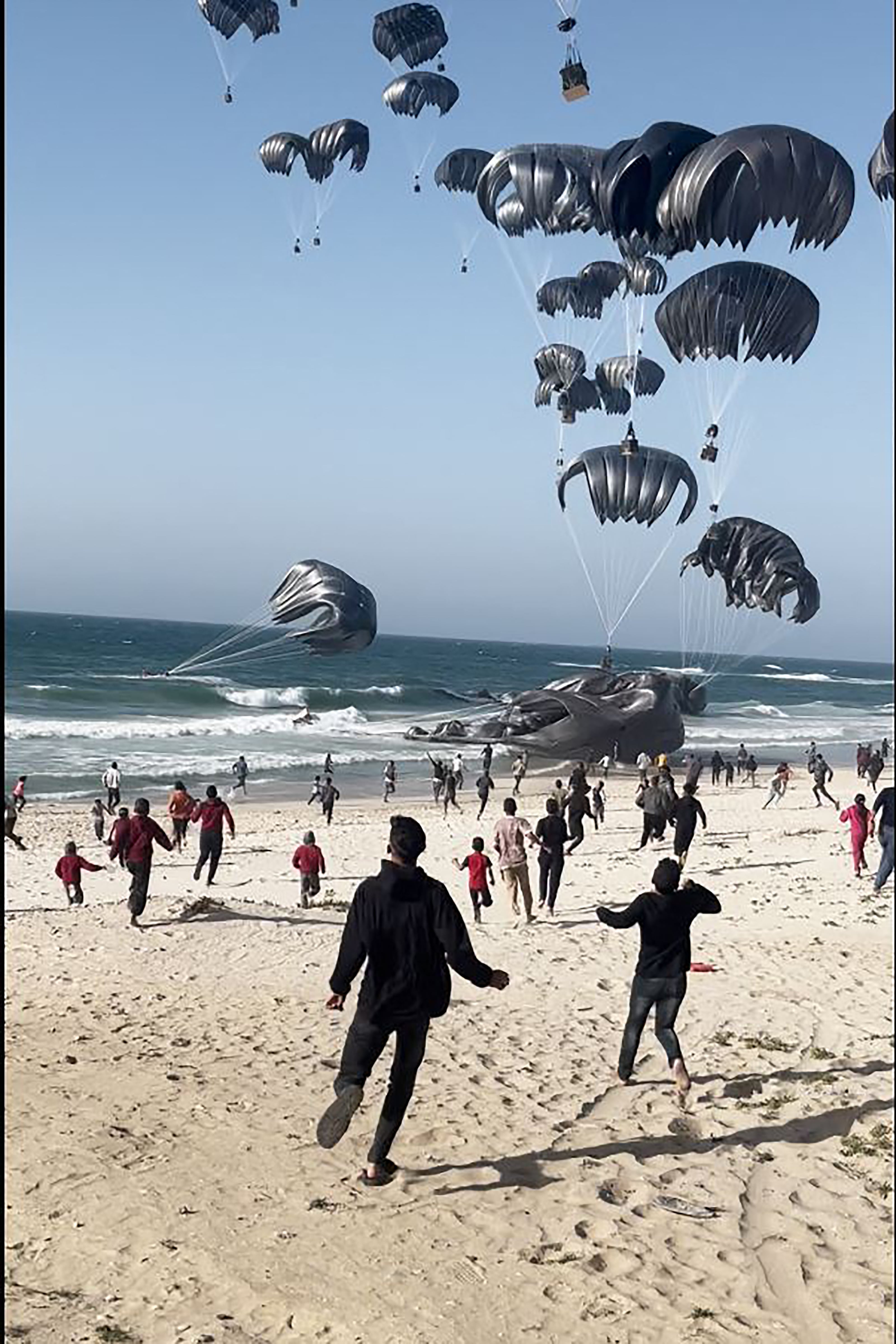 Palestinos corriendo hacia paracaídas sujetos a paquetes de alimentos lanzados desde aviones estadounidenses en una playa en la Franja de Gaza el 2 de marzo de 2024. (Foto de AFP).