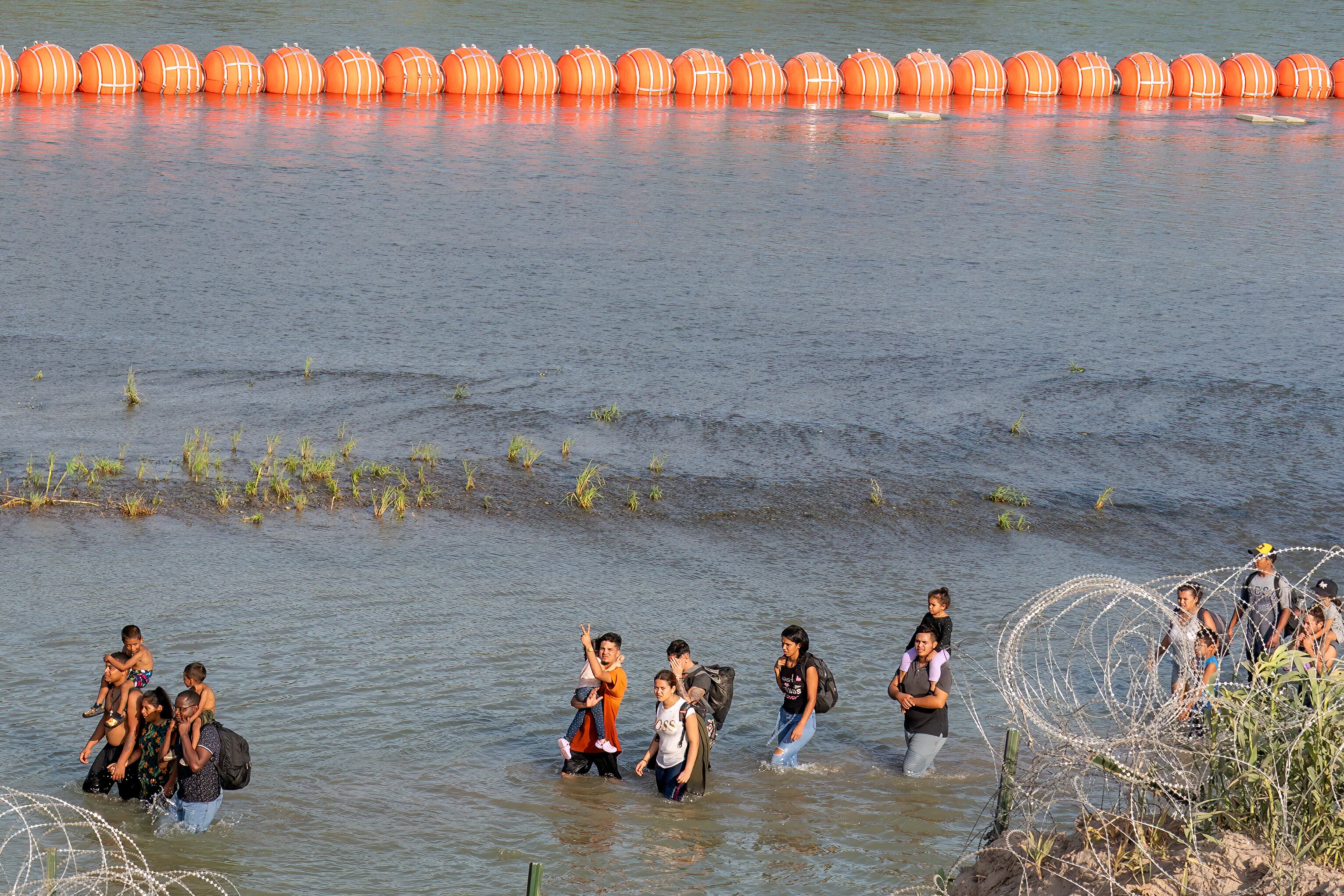 Migrantes caminan entre el alambre y una serie de boyas colocadas en el agua a lo largo de la frontera del Río Grande con México en Eagle Pass, Texas, el 16 de julio de 2023. (Foto de SUZANNE CORDEIRO / AFP).
