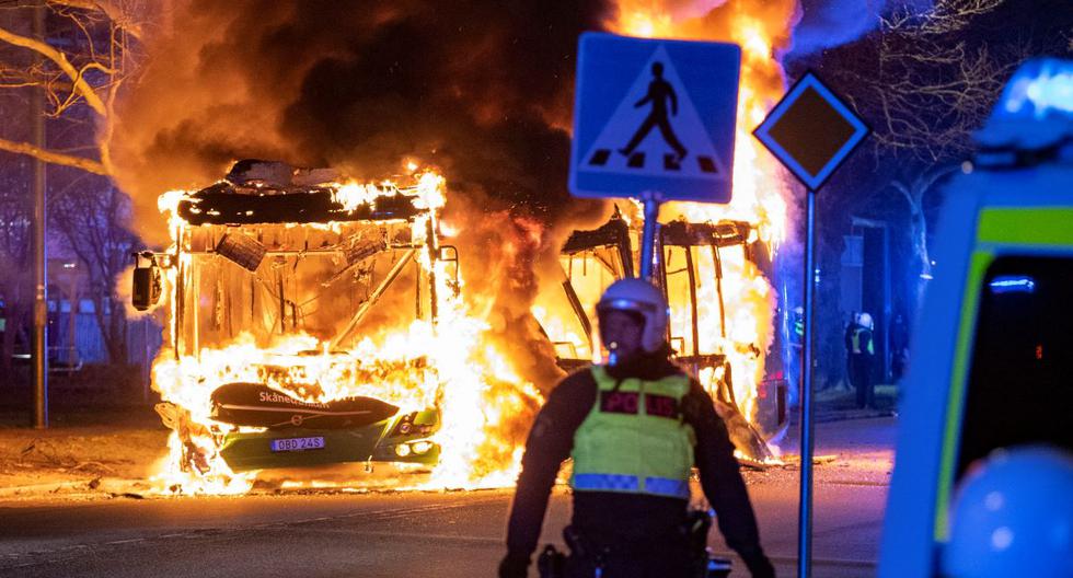 Un oficial de la policía antidisturbios se encuentra junto a un autobús urbano en llamas en Malmo, Suecia, en la noche del 16 de abril de 2022. (Johan NILSSON / AGENCIA DE NOTICIAS TT / AFP).