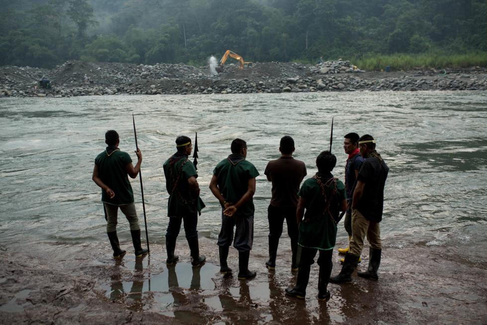 Miembros de la comunidad indígena Cofán de Sinangoe caminan a orillas del río Aguarico, mientras al fondo se ve una retroexcavadora removiendo tierra.